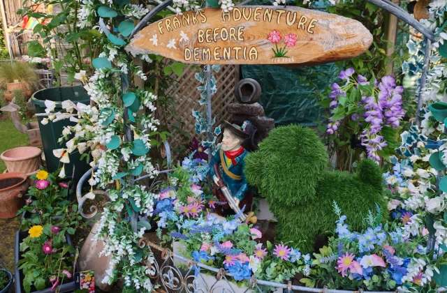 A whimsical garden display featuring a statue, a faux dog, and flowers beneath a sign reading "Frank's Adventure Before Dementia."