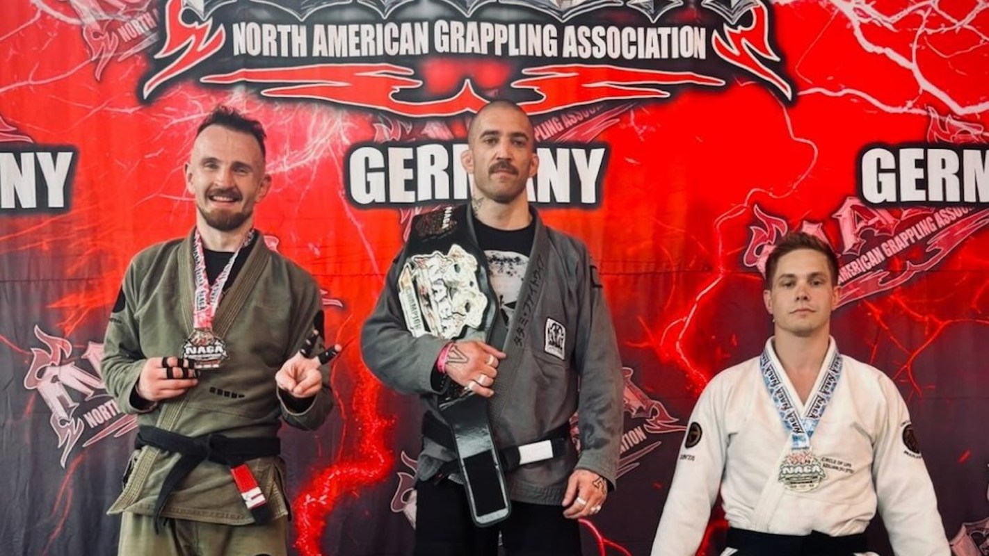 Three grapplers pose with medals and a championship belt in front of a vibrant banner for the North American Grappling Association.