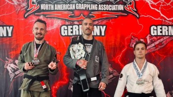 Three grapplers pose with medals and a championship belt in front of a vibrant banner for the North American Grappling Association.