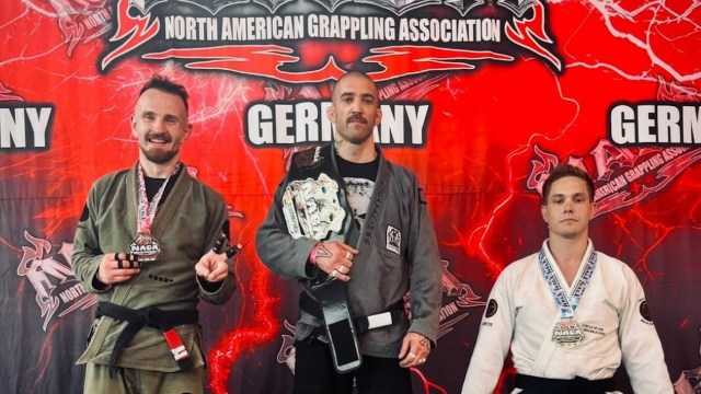 Three grapplers pose with medals and a championship belt in front of a vibrant banner for the North American Grappling Association.