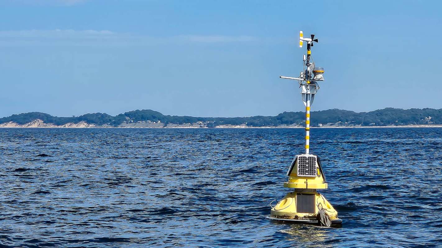 A yellow buoy with sensors and a wind vane floats on blue water, with a distant landmass in the background.