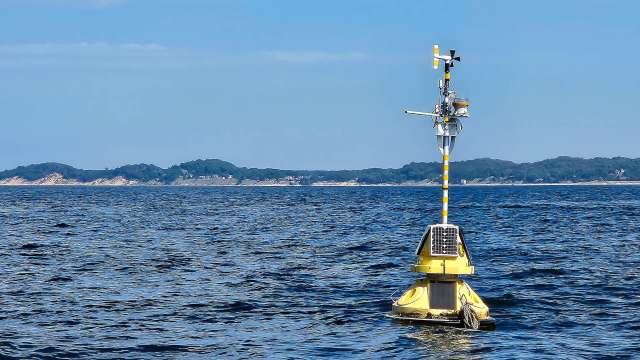 A yellow buoy with sensors and a wind vane floats on blue water, with a distant landmass in the background.