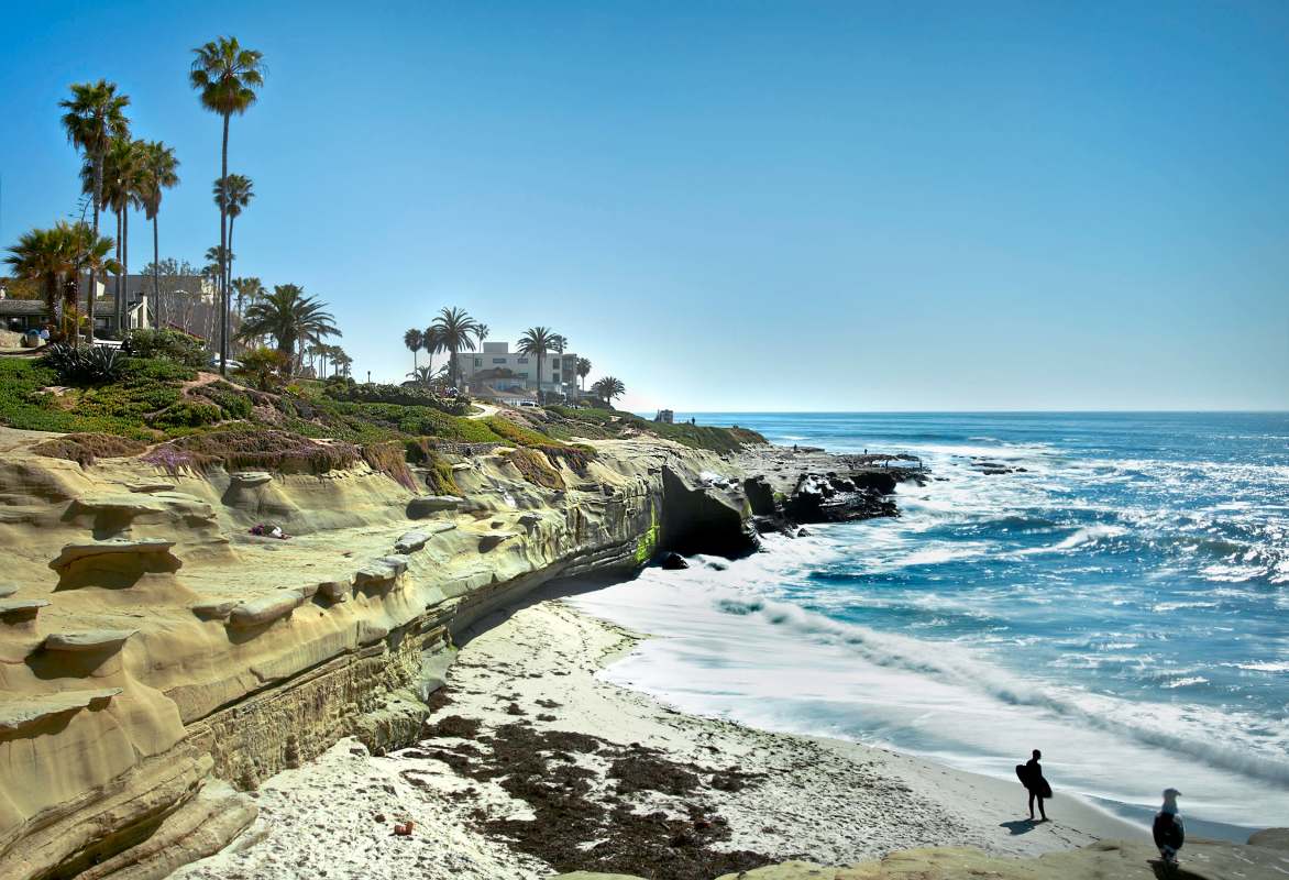A coastal scene featuring cliffs, palm trees, and people enjoying the beach with waves crashing nearby.