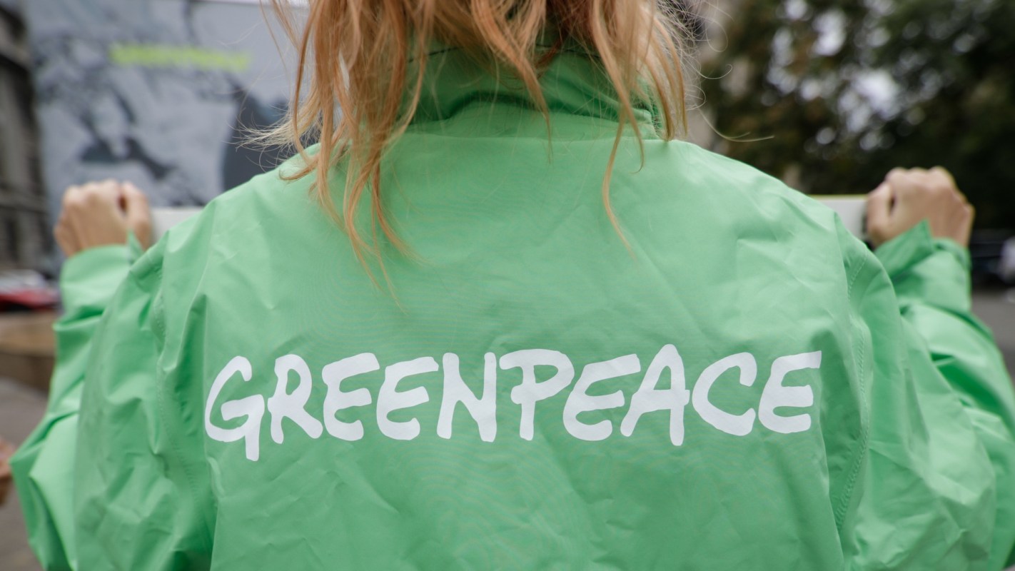 A person wearing a green jacket with "GREENPEACE" printed on the back stands outdoors.