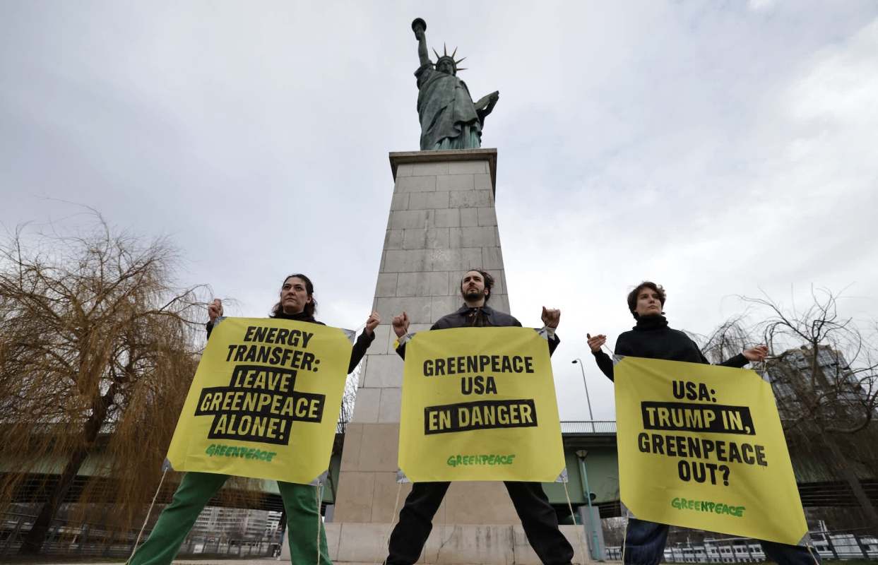 Three protesters hold large yellow signs with messages about Greenpeace in front of the Statue of Liberty.