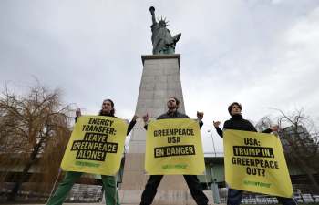 Three protesters hold large yellow signs with messages about Greenpeace in front of the Statue of Liberty.