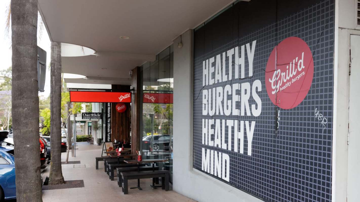 A sidewalk view of a Grill'd restaurant featuring a large sign promoting healthy burgers and outdoor seating.
