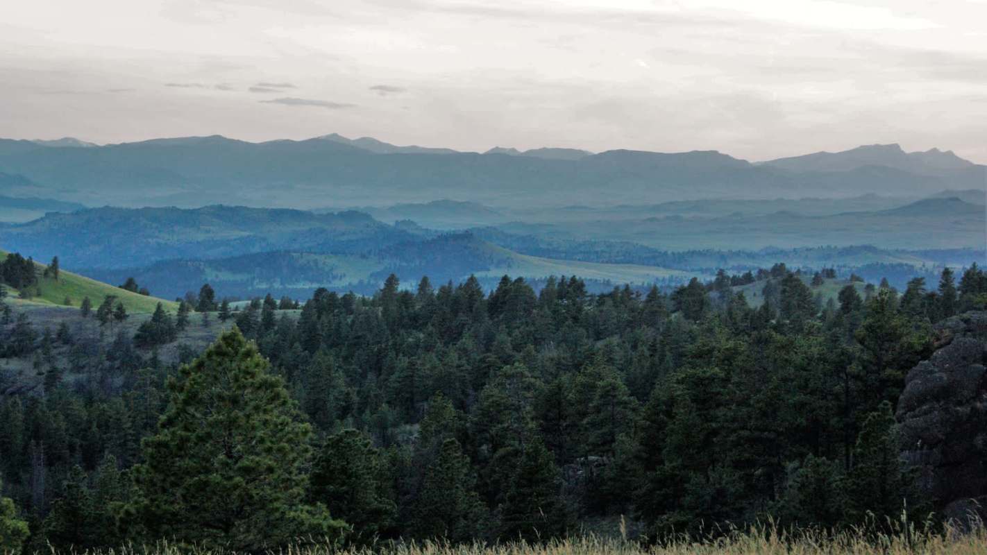 A panoramic view of rolling green hills and layered mountains under a cloudy sky at dusk.
