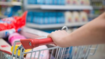 A hand grips the handle of a shopping cart filled with various grocery items in a store aisle.