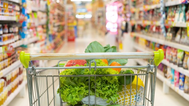 A grocery cart filled with fresh vegetables and fruit in a brightly lit supermarket aisle.
