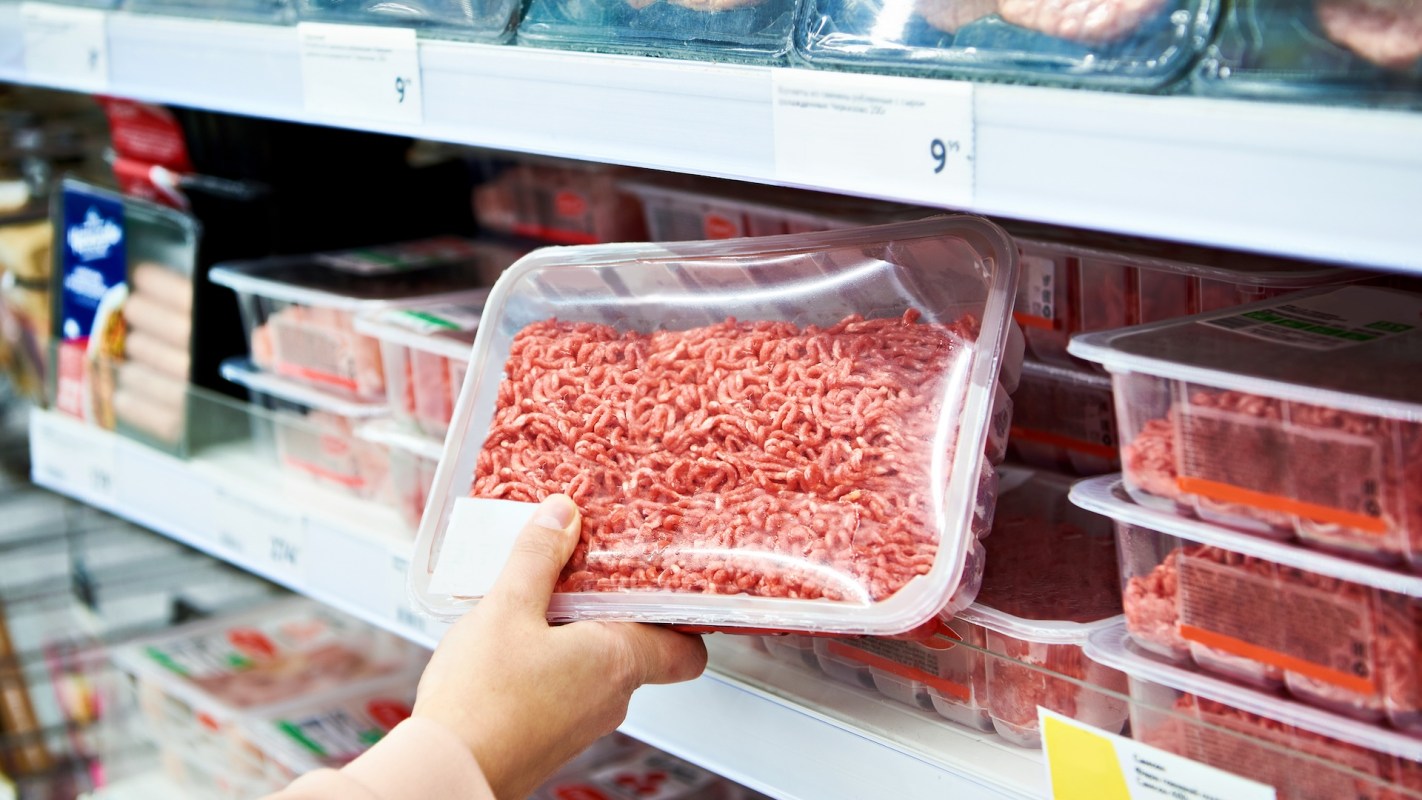 A person holding a package of ground meat in a grocery store's refrigerated section.