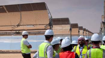 A group of professionals in hard hats and vests discuss solar panels in a desert setting.