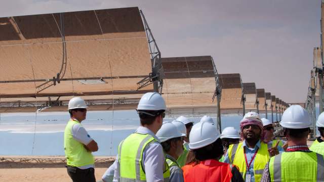 A group of professionals in hard hats and vests discuss solar panels in a desert setting.