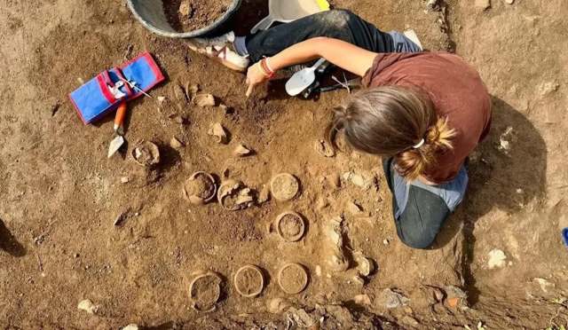 An archaeologist digs in the soil, revealing pottery fragments and artifacts on a site.