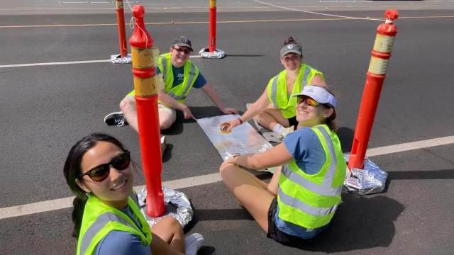 A group of four people wearing neon vests sits on the road, surrounded by traffic cones, smiling and engaging in activities.