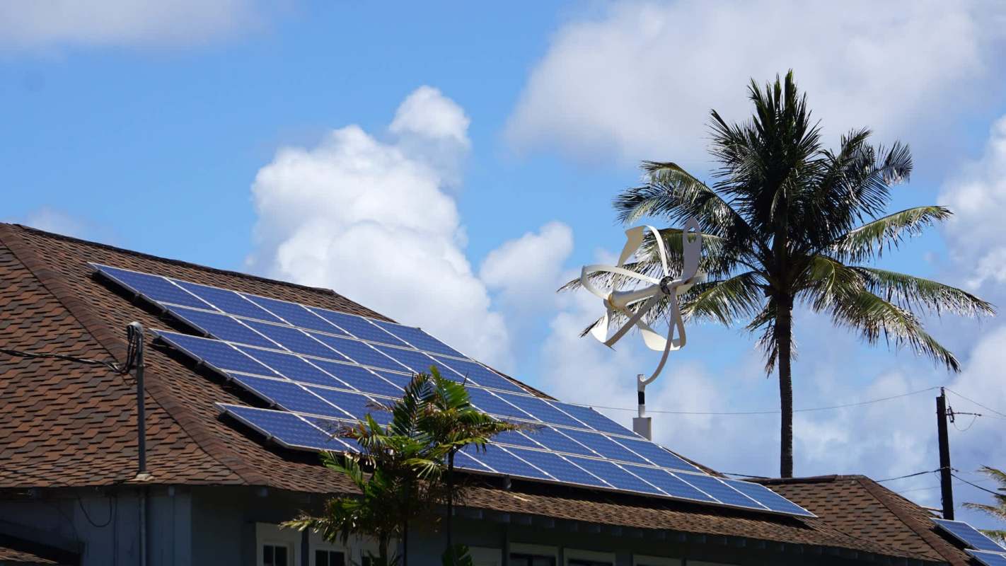 A rooftop equipped with solar panels and a small wind turbine, surrounded by palm trees and blue sky.