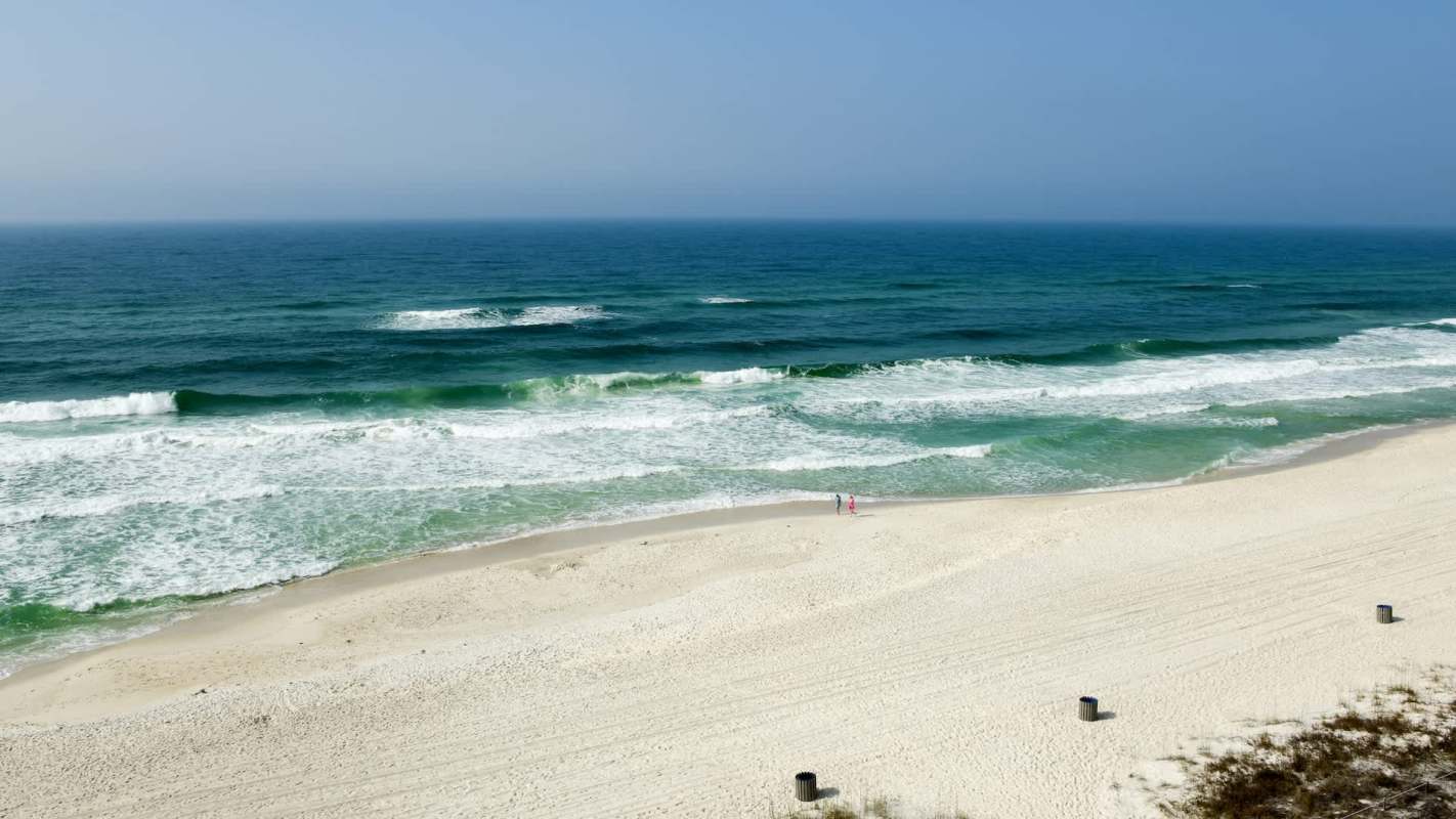 A couple walks along a sandy beach with rolling waves and a clear blue sky in the background.