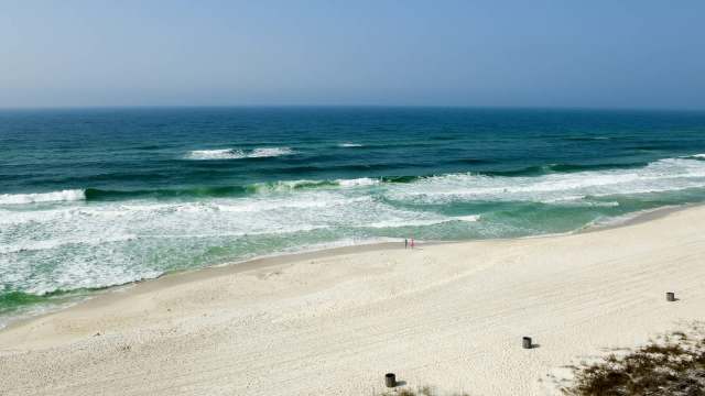 A couple walks along a sandy beach with rolling waves and a clear blue sky in the background.