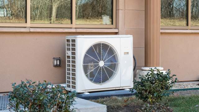 A heat pump unit installed next to a building, surrounded by small shrubs and grass.