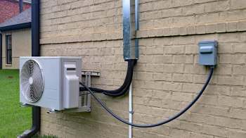An outdoor heat pump unit mounted on a wall, with electrical and drainage connections visible.