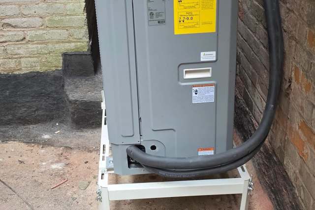 An HVAC unit on a snow stand near a brick wall.