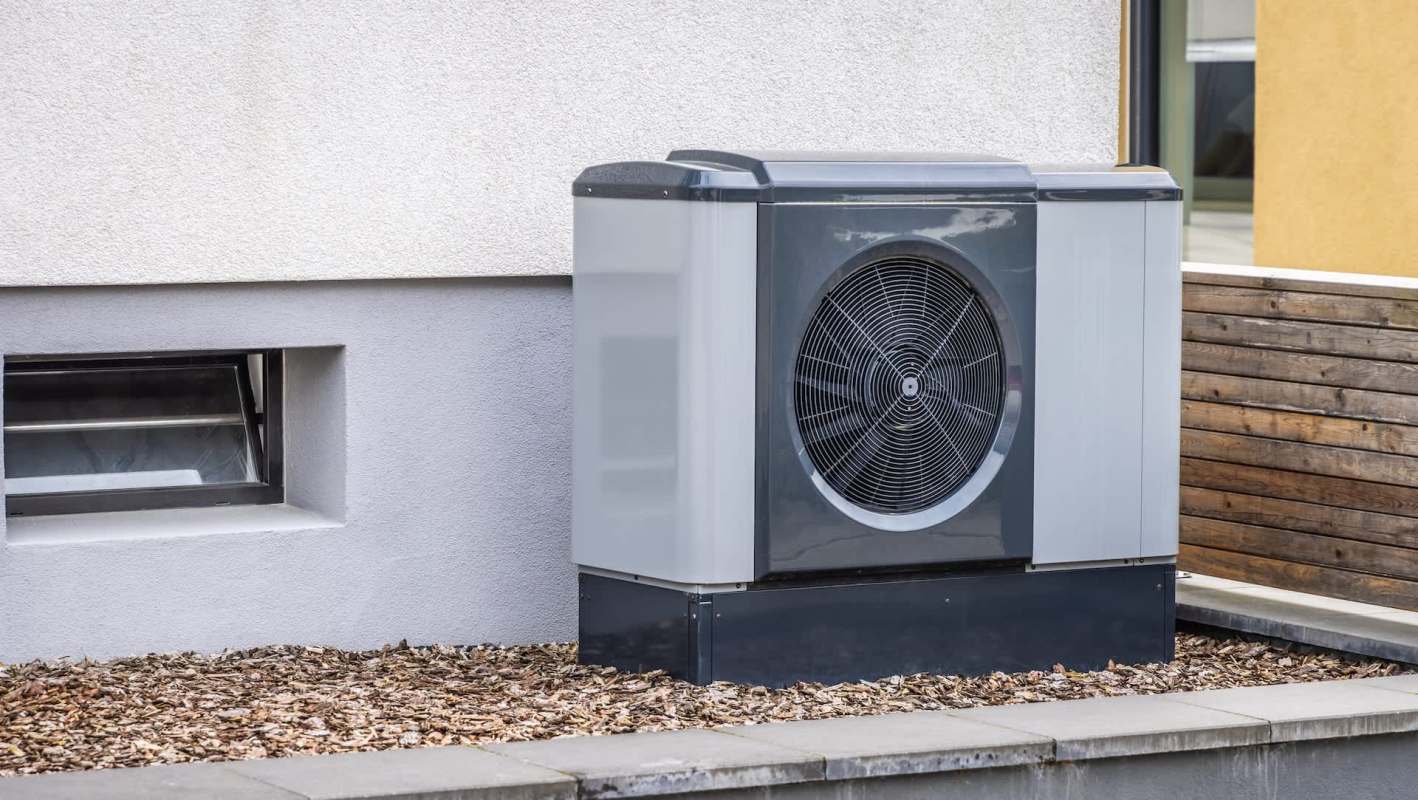 A modern heat pump unit placed beside a building's exterior wall with mulch landscaping.