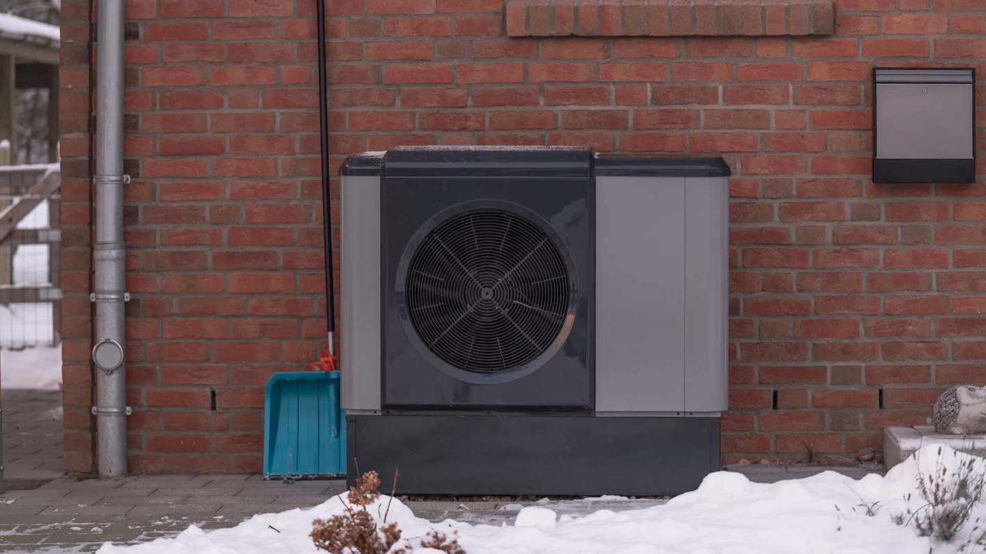 A heat pump outside of a brick house with a shovel next to it and snow on the ground.