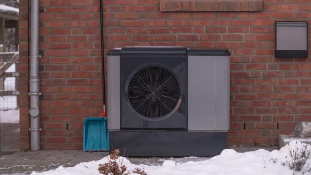 A heat pump outside of a brick house with a shovel next to it and snow on the ground.