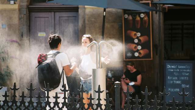 Two people converse outside a cafe while being sprayed by a misting machine.