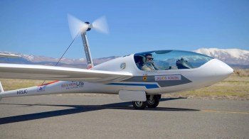 A sleek experimental aircraft with a propeller, taxiing on a runway against a clear blue sky and distant mountains.