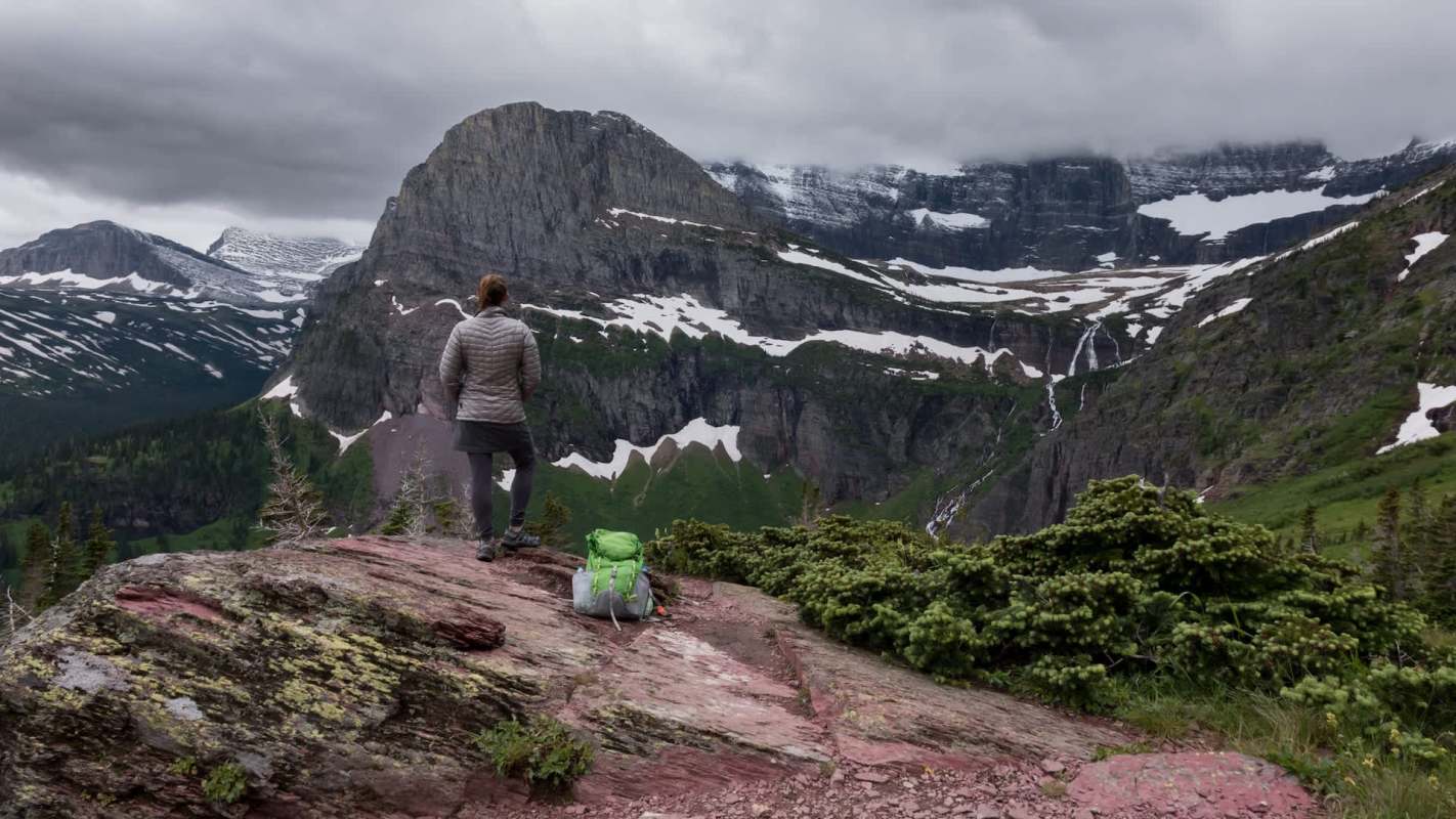 A hiker stands on a rocky outcrop overlooking a mountain landscape with snow and greenery under a cloudy sky.