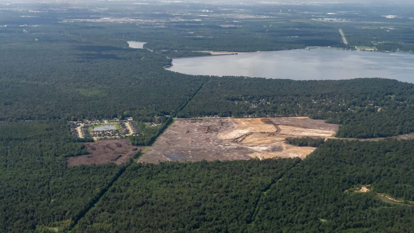 An aerial view shows a cleared area for a construction site among dense trees near a lake and a residential neighborhood.