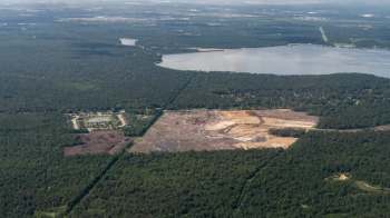 An aerial view shows a cleared area for a construction site among dense trees near a lake and a residential neighborhood.