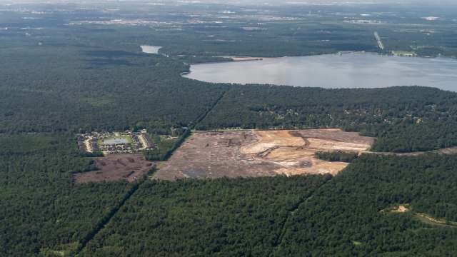 An aerial view shows a cleared area for a construction site among dense trees near a lake and a residential neighborhood.