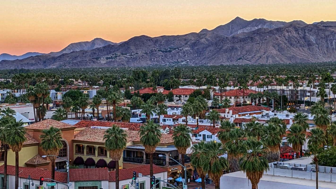 Aerial view of a palm tree-lined town with red-roofed buildings and mountains in the background at sunset.