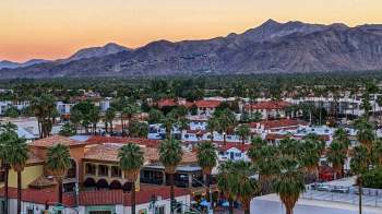 Aerial view of a palm tree-lined town with red-roofed buildings and mountains in the background at sunset.