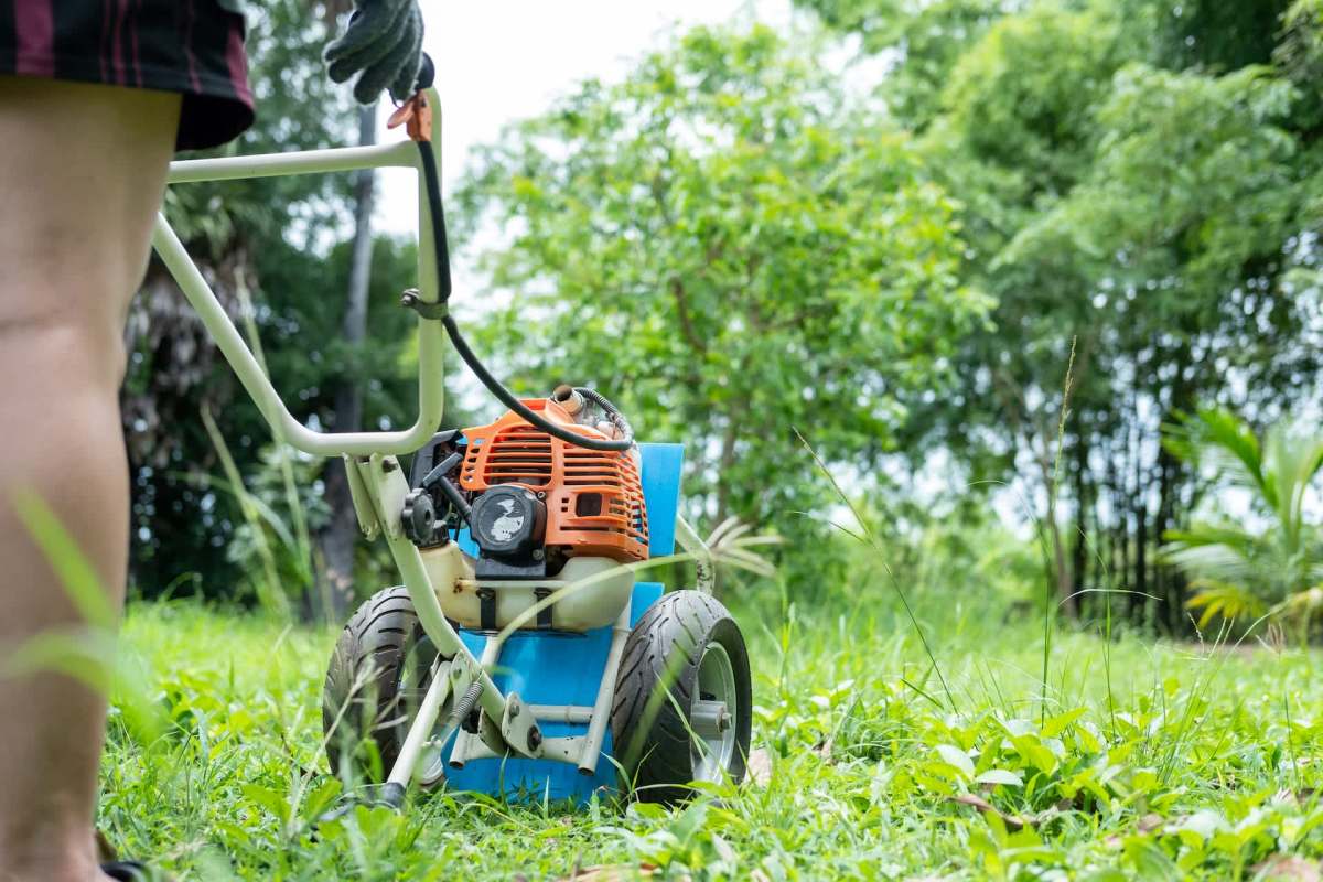 A person prepares to use a gas-powered tiller in a lush, green garden surrounded by trees.