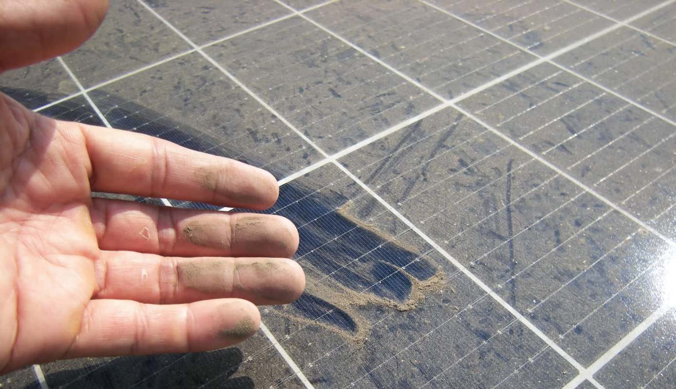 A hand with dirt on the fingers is positioned over a dusty solar panel surface.