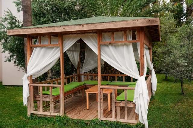 A backyard wooden gazebo with green cushions and sheer white curtains.