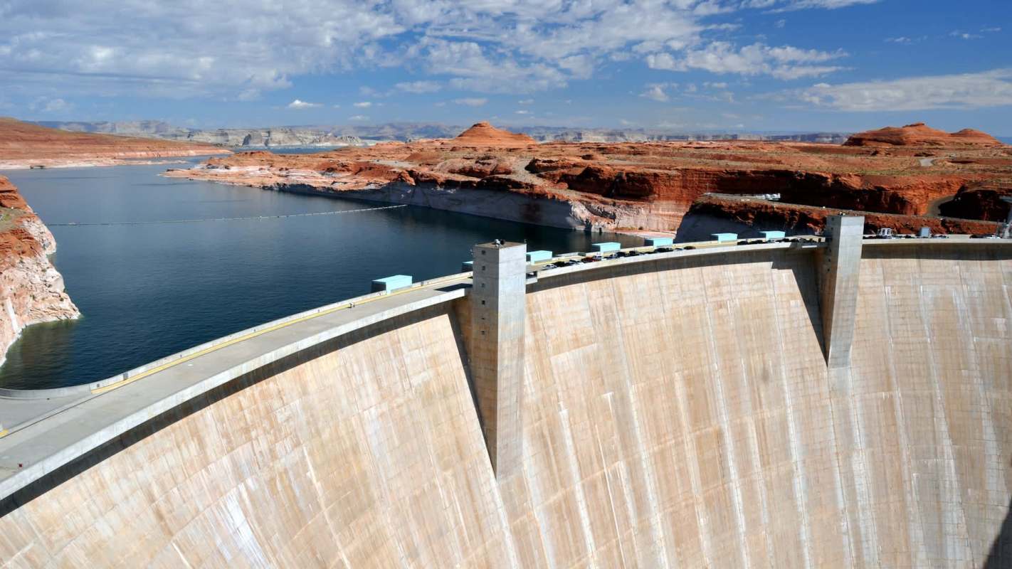 A view of a large dam beside a lake, surrounded by red rock formations and under a bright blue sky.