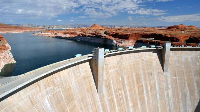 A view of a large dam beside a lake, surrounded by red rock formations and under a bright blue sky.