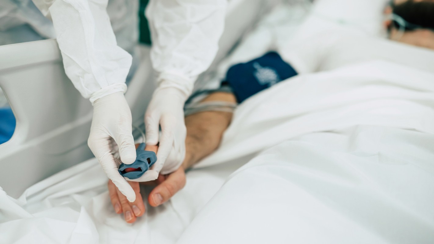 A healthcare worker in gloves fits a pulse oximeter on a patient's finger in a hospital setting.