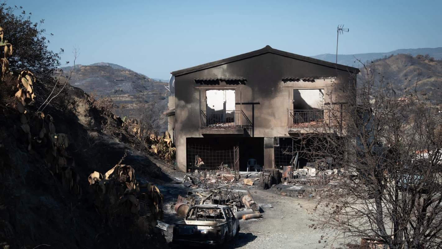 A burned-out house surrounded by charred landscape and debris from recent wildfires.