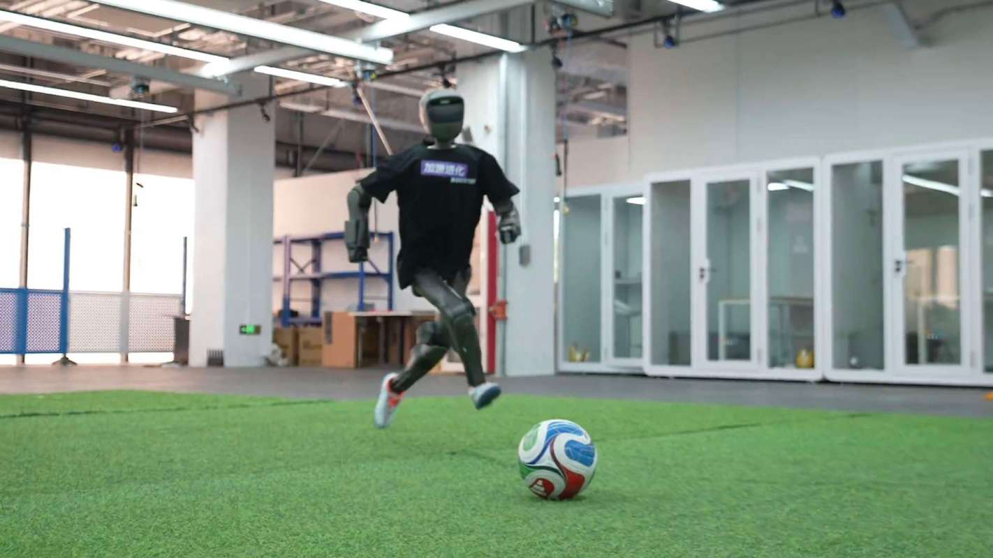 A humanoid robot in a t-shirt runs towards a soccer ball on artificial grass indoors.