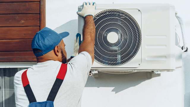 A technician in gloves installs an outdoor air conditioning unit on a wall.