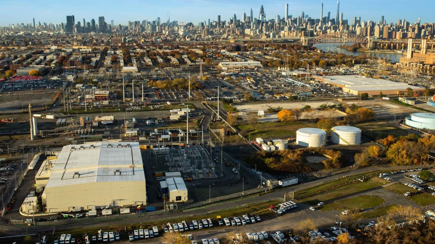 Aerial view of an industrial area with warehouses, utility structures, and the New York City skyline in the background.