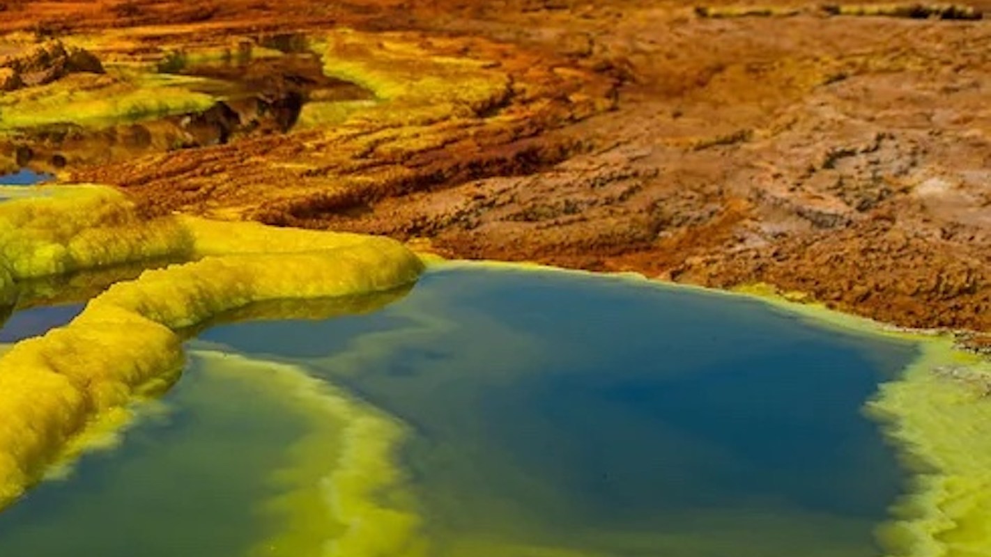 Colorful geothermal landscape with yellow and blue mineral pools surrounded by textured, rust-colored earth.