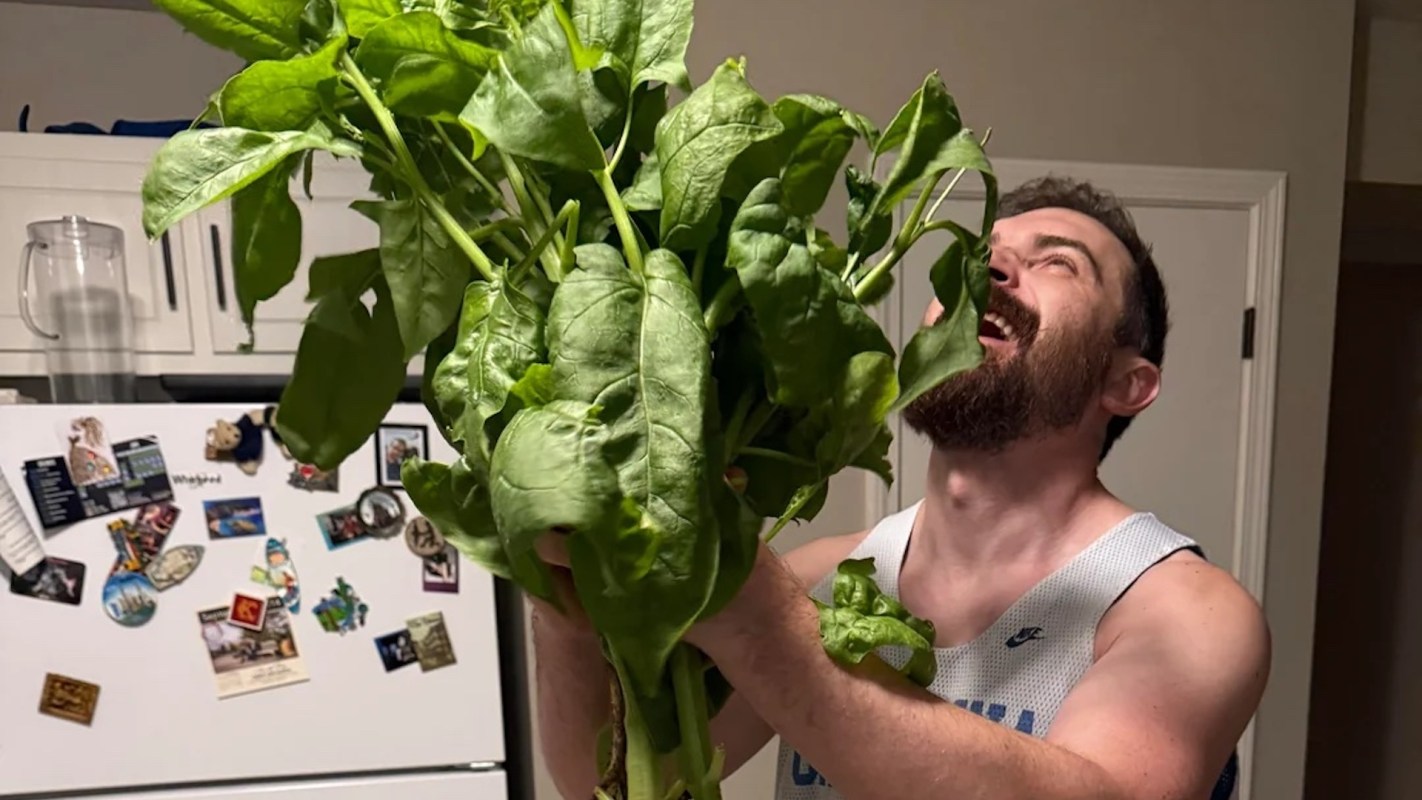 A man joyfully holds a large bunch of fresh spinach in a kitchen.