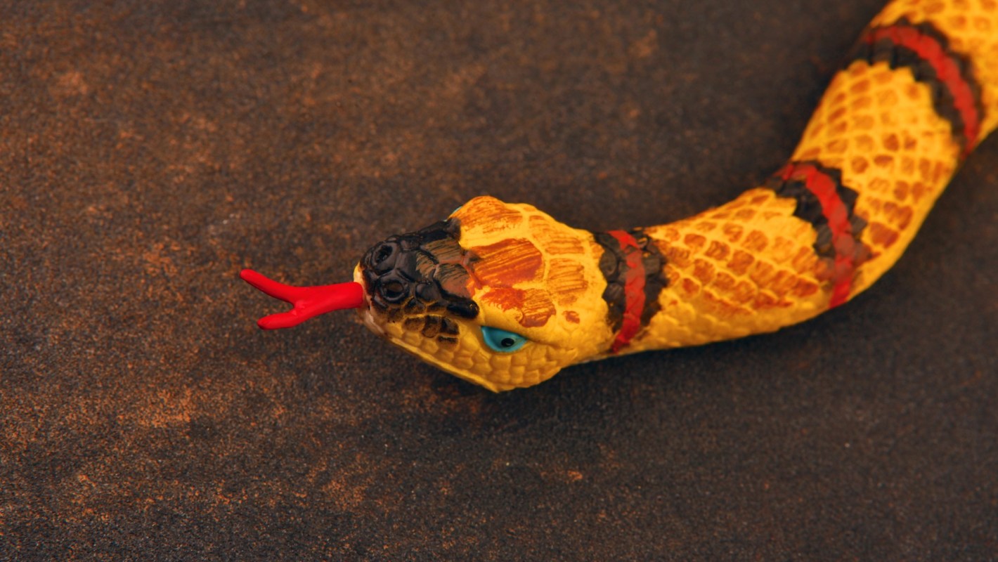 A close-up of a toy snake with yellow scales and red markings, tongue flicking out against a dark background.