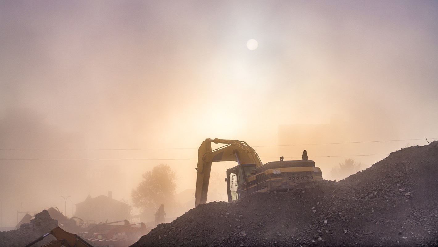 A construction site shrouded in fog with an excavator silhouette against a hazy sunrise.
